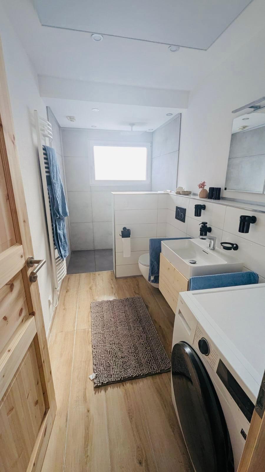 Badezimmer in einer Ferienwohnung mit Waschtrockner und begehbarer Dusche. Mit Holzoptik Fliesen und Tageslicht mit Milchglasfenster