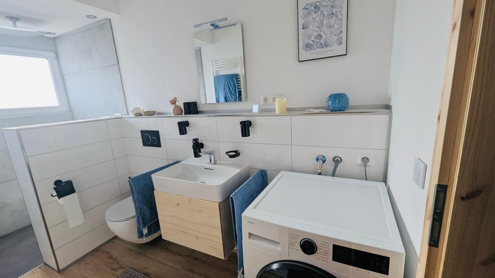 Badezimmer in einer Ferienwohnung mit Waschtrockner und begehbarer Dusche. Mit Holzoptik Fliesen und Tageslicht mit Milchglasfenster